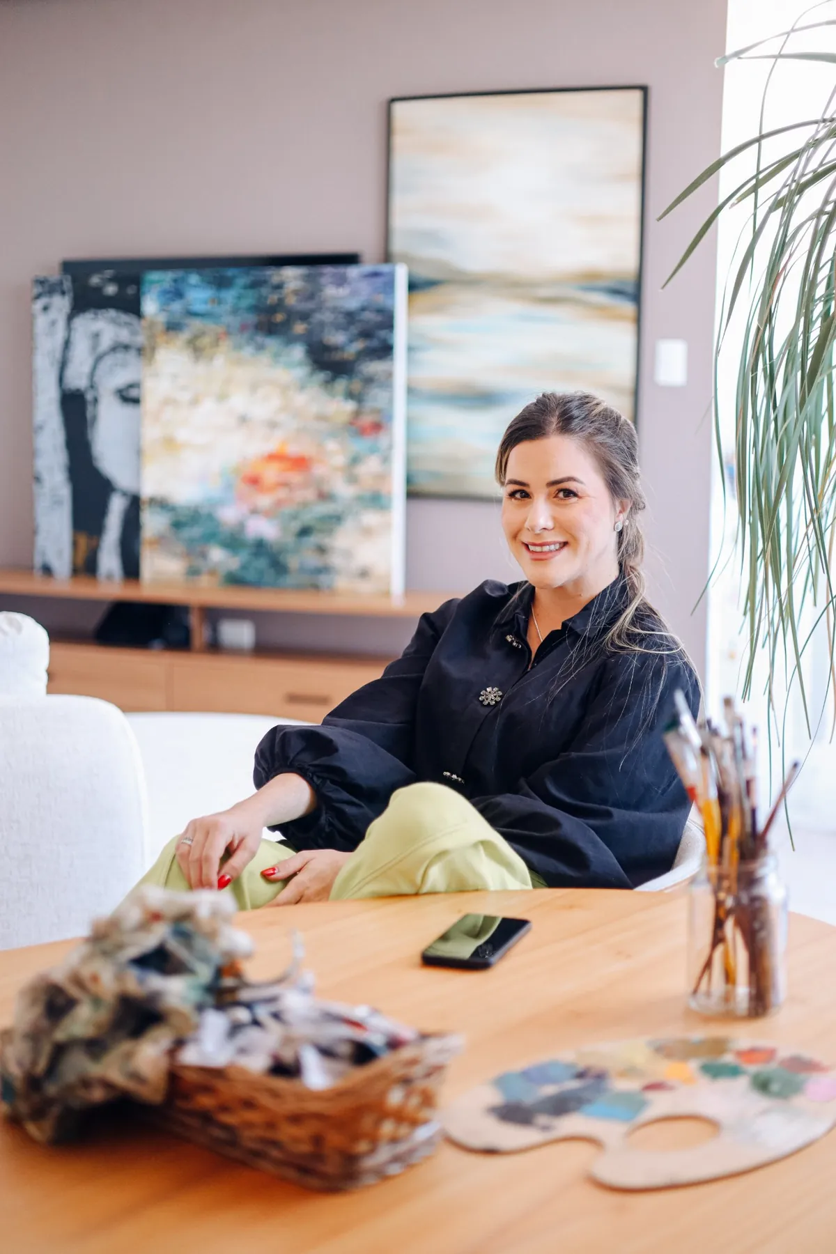 Michele Anadão sorrindo, sentada à mesa com pincéis e uma paleta de tintas, com quadros ao fundo